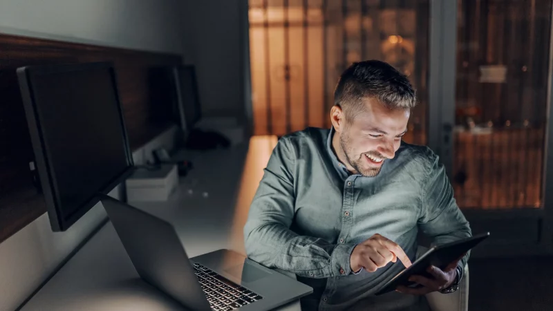 Bärtiger Freiberufler mit zahnigem Lächeln, der spät abends im Büro sitzt und ein Tablet benutzt. Neben ihm auf dem Schreibtisch ein Laptop