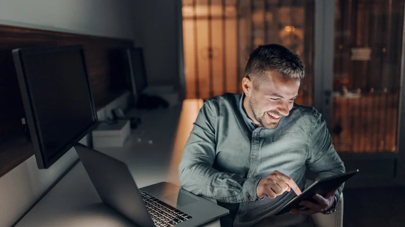 Bärtiger Freiberufler mit zahnigem Lächeln, der spät abends im Büro sitzt und ein Tablet benutzt. Neben ihm auf dem Schreibtisch ein Laptop