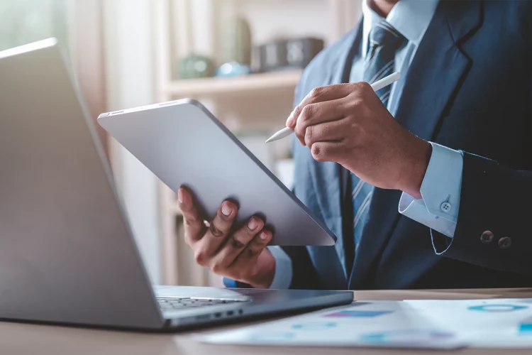 Mann schreibt mit einem Stift auf ein Tablet und sitzt dabei vor einem Laptop