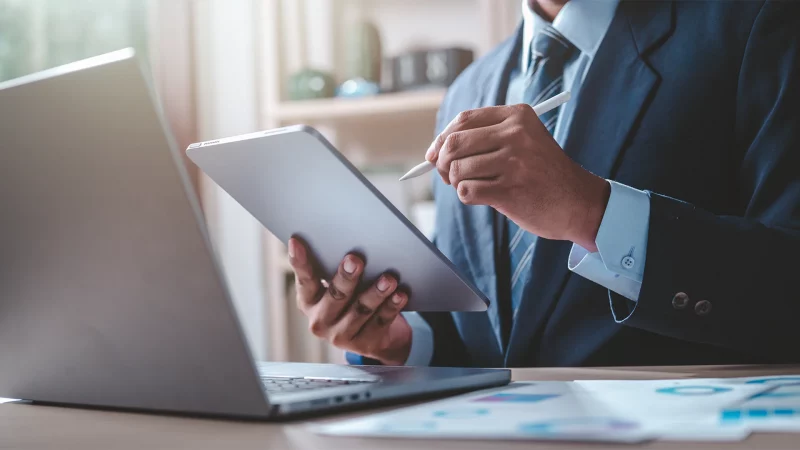 Mann schreibt mit einem Stift auf ein Tablet und sitzt dabei vor einem Laptop