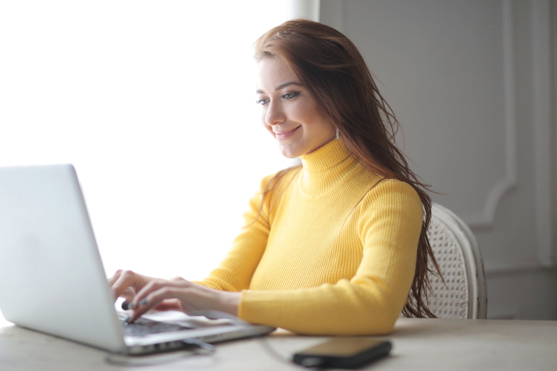 Eine Frau mit gelbem Pullover sitzt lächelnd am Laptop und schreibt